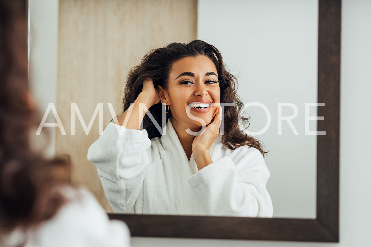 Beautiful smiling brunette in a white bathrobe looking at her reflection in the bathroom mirror	