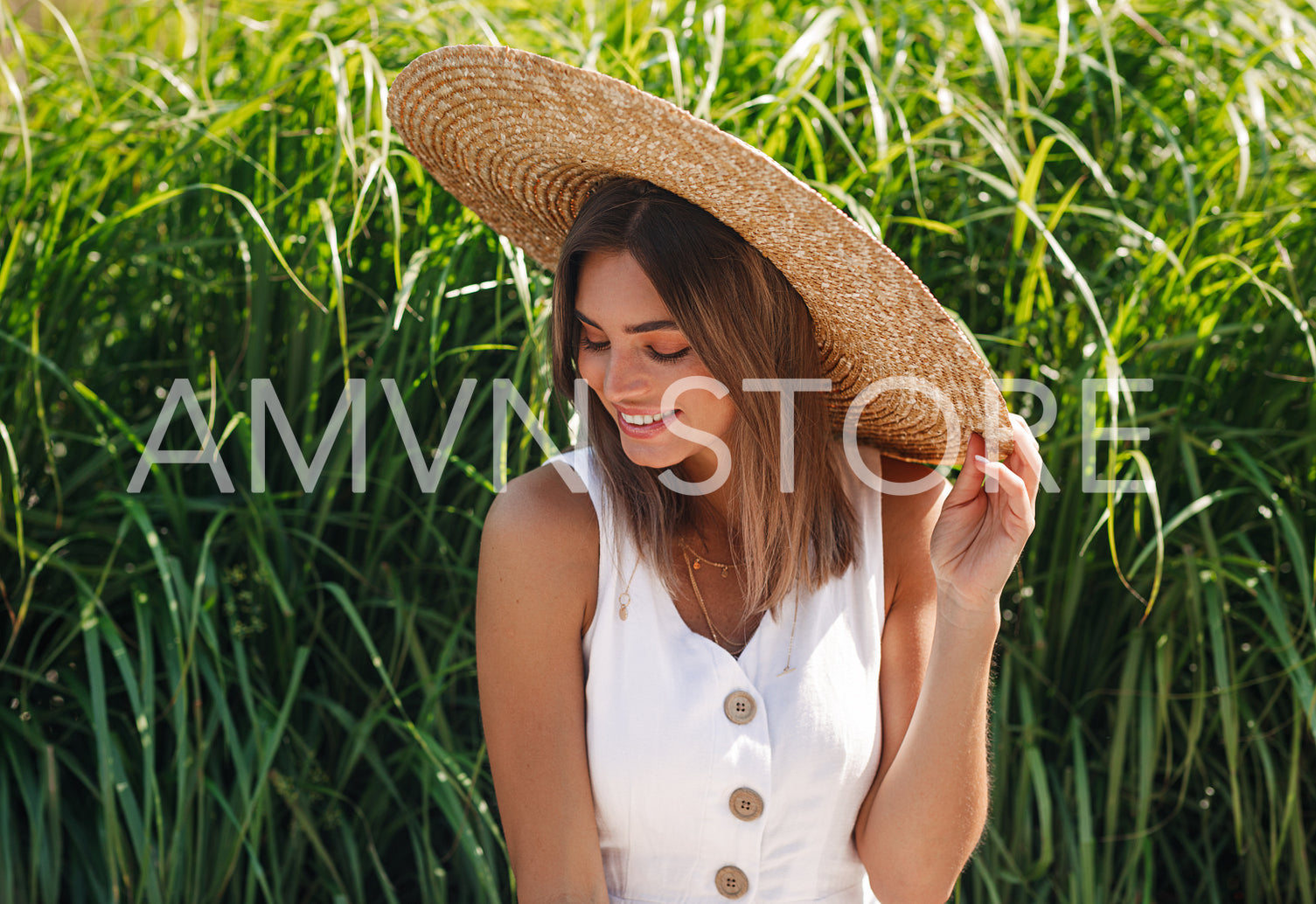 Happy stylish woman sitting in front of grass and looking while holding her hat	