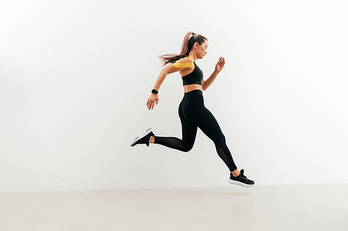 Runner in sport clothes sprinting near a white wall. Woman with kinesiology tape on shoulder jogging indoors.