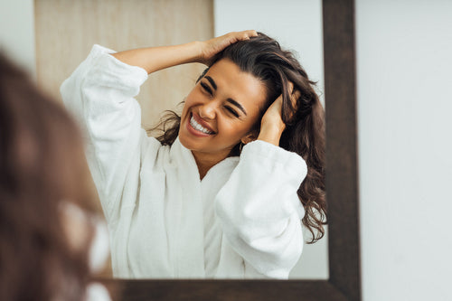 Happy brunette woman with palms in her hair looking at mirror reflection in the bathroom