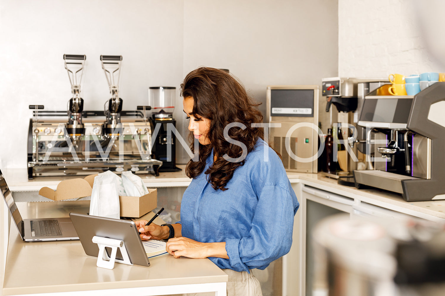 Side view of businesswoman in her cafe verifying the invoice before shipping the order. Woman working at desk with laptop and box with packs of coffee.	