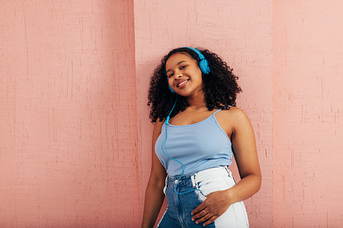 Brunette woman with curly hait leaning a pink wall while listening music by blue headphones