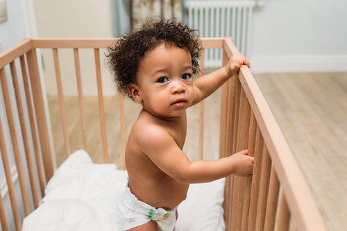 Portrait of curly haired toddler standing in a crib, looking at camera