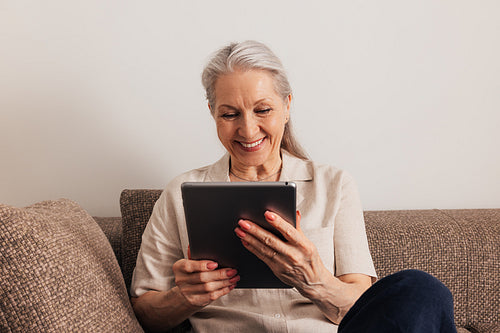 Close up of a smiling senior woman with grey hair holding digital tablet while sitting on couch