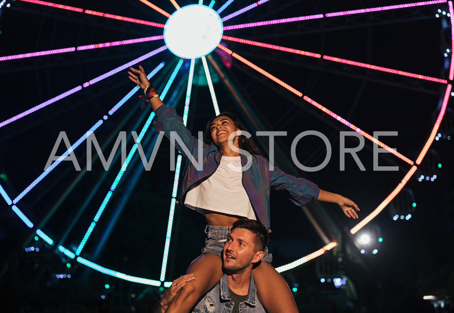 Happy woman sitting on the shoulders of her boyfriend and having fun. Young couple against ferris wheel during the festival.