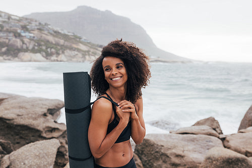 Portrait of a beautiful smiling woman standing with yoga mat by seaside. Female in sportswear relaxing after yoga looking away.