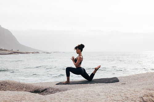 Side view of fit woman practicing yoga on mat by ocean. Female meditating at evening with folded hands.