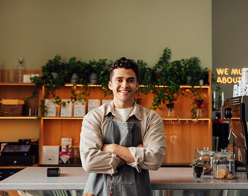 Coffee shop owner in an apron standing with crossed arms and smiling. Young handsome man leaning counter looking at a camera in a coffee shop.