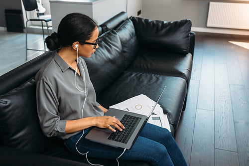 Young woman sitting in a living room in her apartment on a couch with laptop and looking on documents