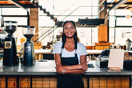 Portrait of a smiling coffee shop owner standing at counter wearing apron and arms crossed