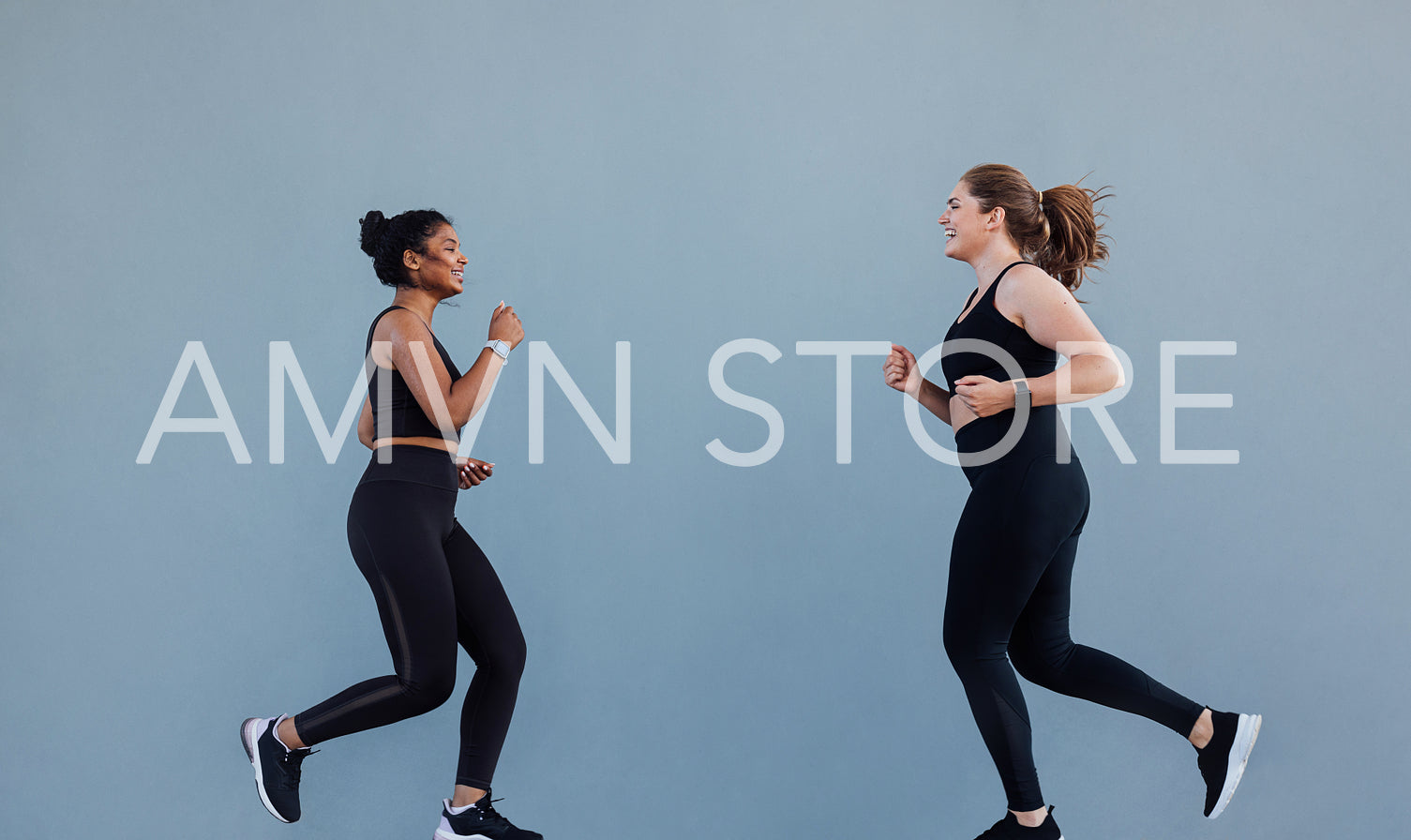 Sportswoman running in front of her friend and motivating to run. Two jogging friends smiling while running at grey wall.