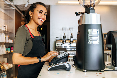 Female barista at counter using cashbox computer in coffee shop. Smiling waitress wearing apron and looking away.