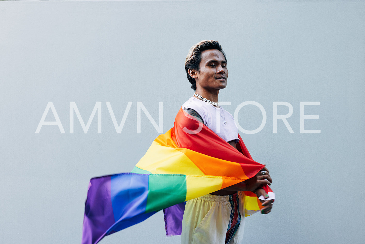 Side view of smiling male with closed eyes holding LGBT rainbow flag against grey wall