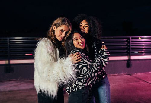 Three beautiful girlfriends embracing each other while standing on roof at neon lights