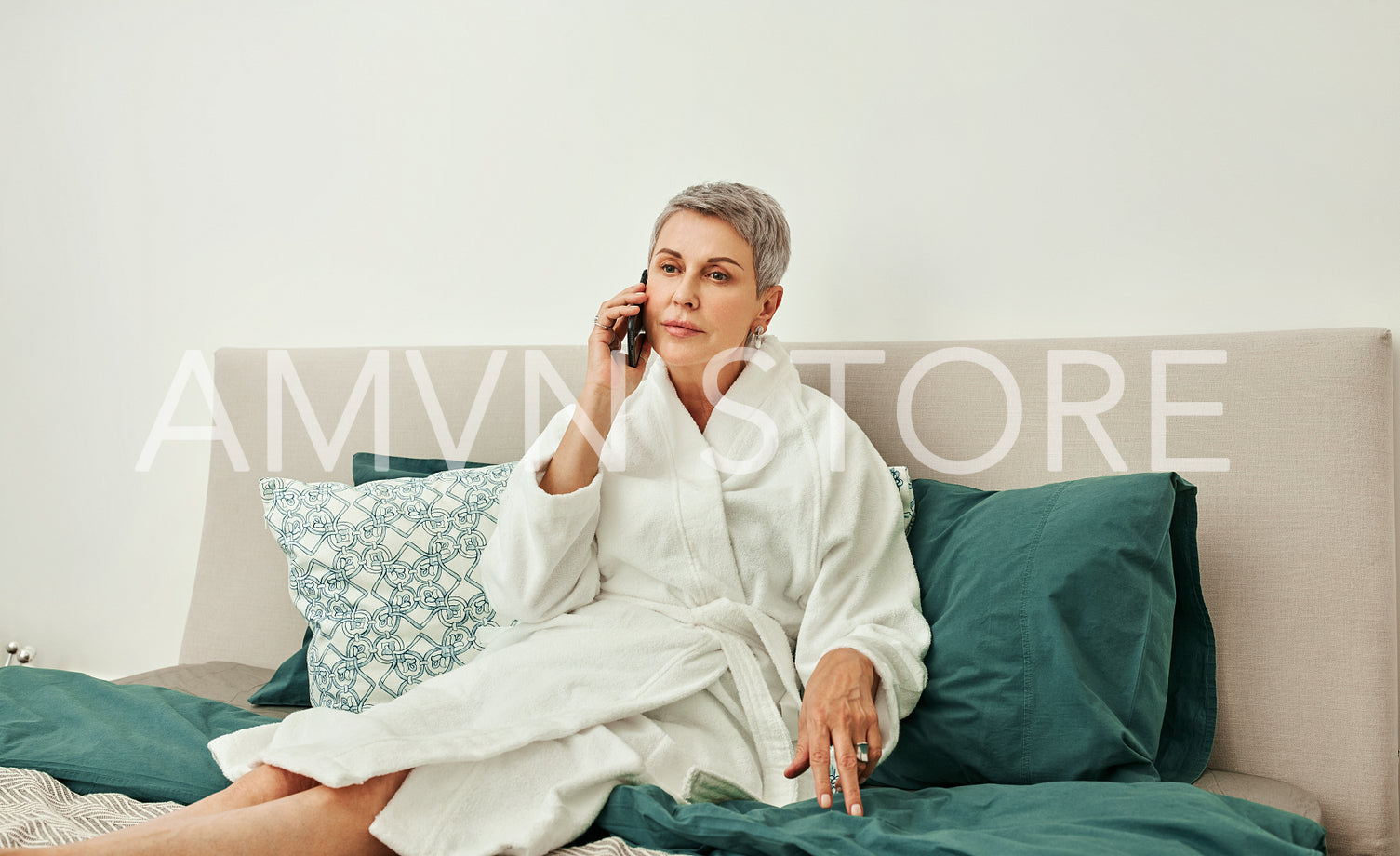 Senior woman with short grey hair talking on a mobile phone from hotel room lying on a bed