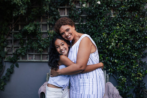 Granny and grandchild embrace and look at the camera. Grandma and granddaughter hugging and smiling while standing together outside.