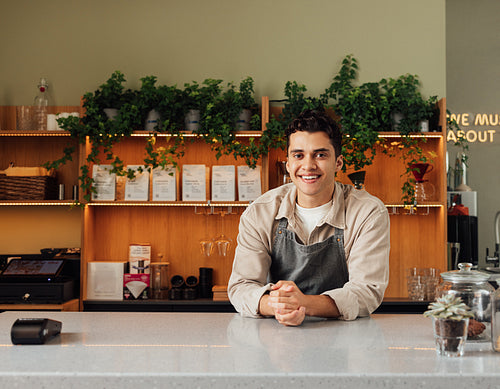 Smiling coffee shop owner in apron leaning a counter. Middle eastern bartender looking at camera.