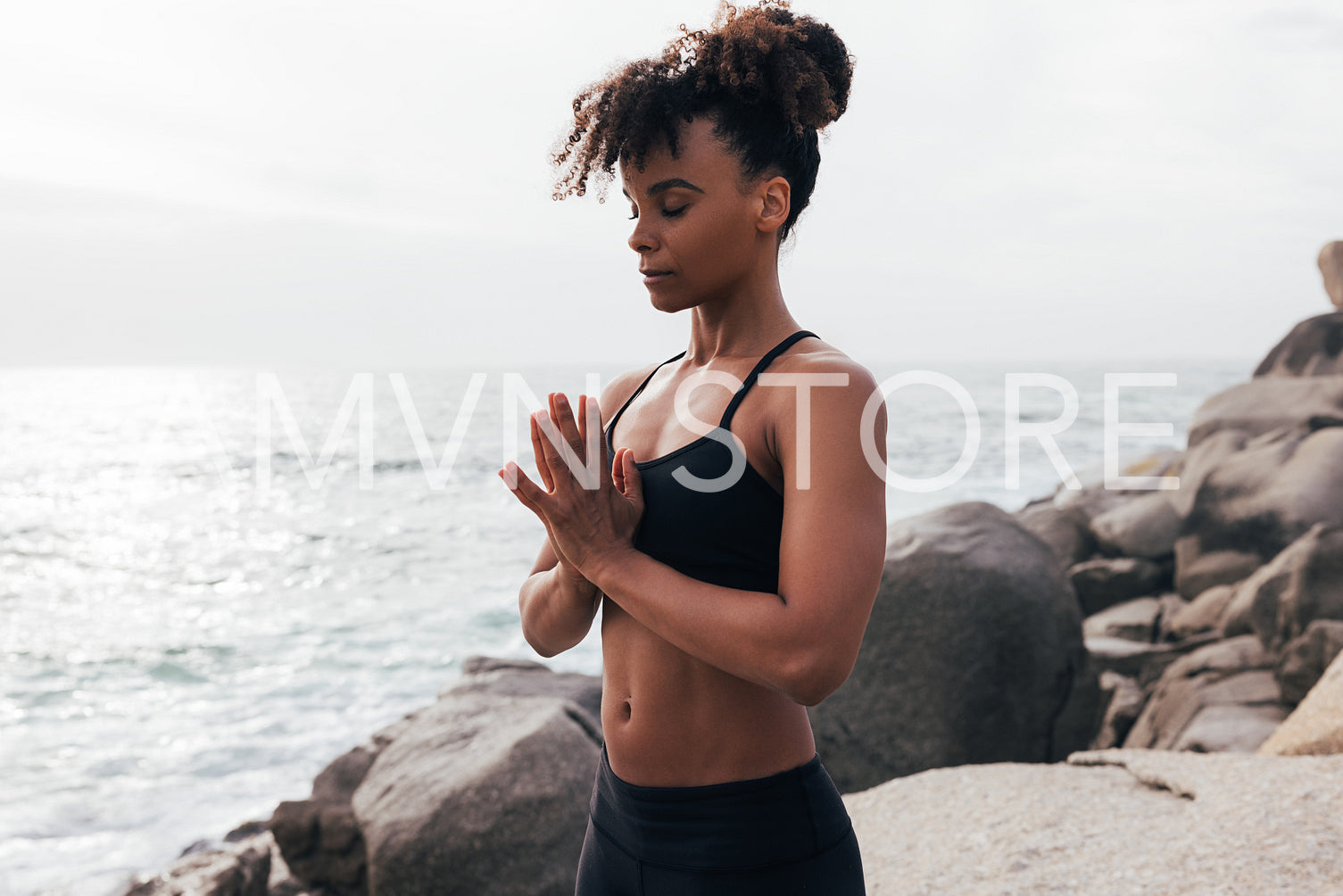 Young woman with folded hands standing at sunset by ocean. Female meditating outdoors with closed eyes.