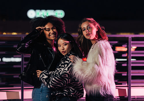 Three stylish girls standing on a street at night in front of neon lights and looking at camera