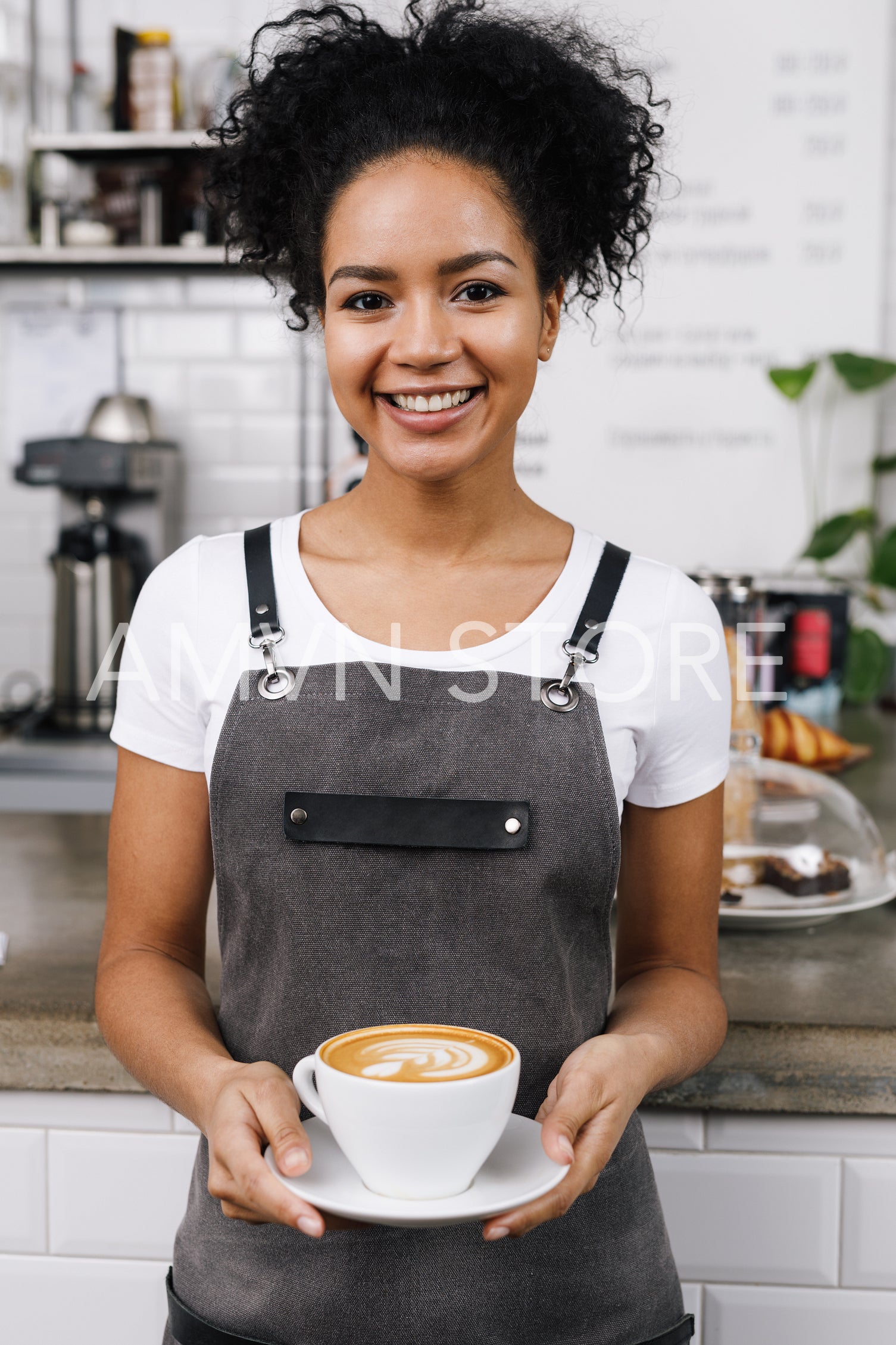 Young smiling waitress holding a cup of latte, standing at bar desk in coffeeshop