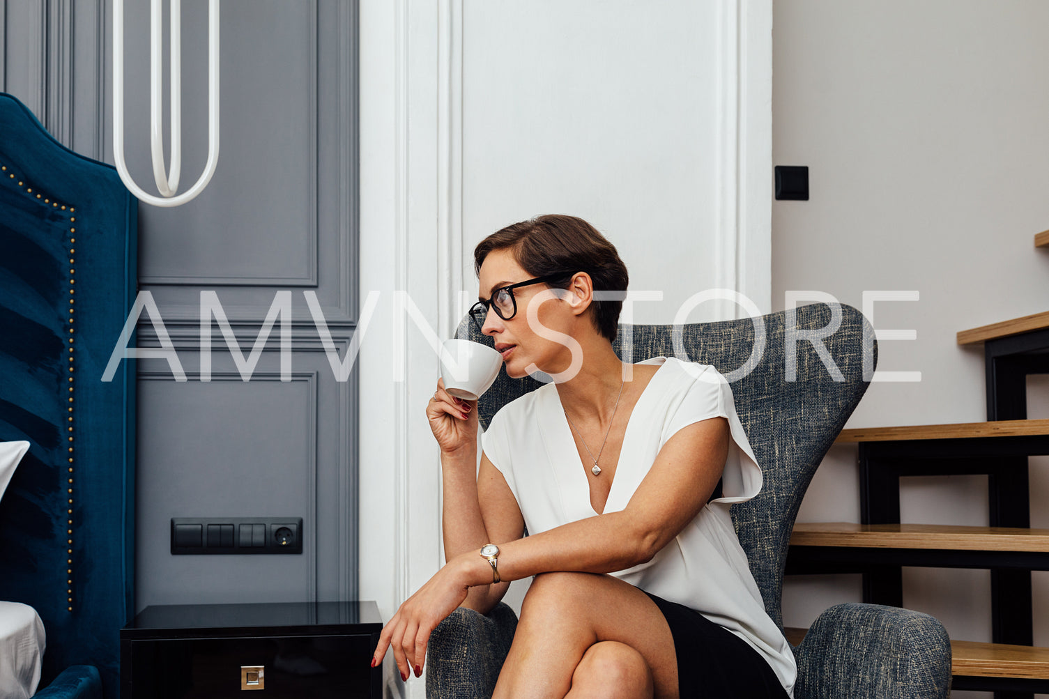 Side view of mid adult woman drinking coffee in a modern apartment. Businesswoman in a hotel room sitting in an armchair and looking away.