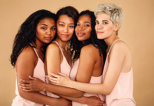 Four beautiful women hugging each other in a studio. Diverse females standing at background.