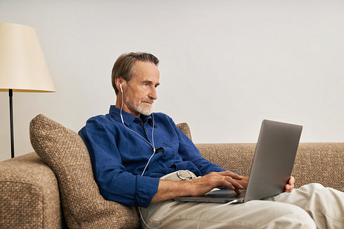 Side view of senior man wearing earphone typing on laptop. Elderly male sitting on sofa with laptop on his hips.