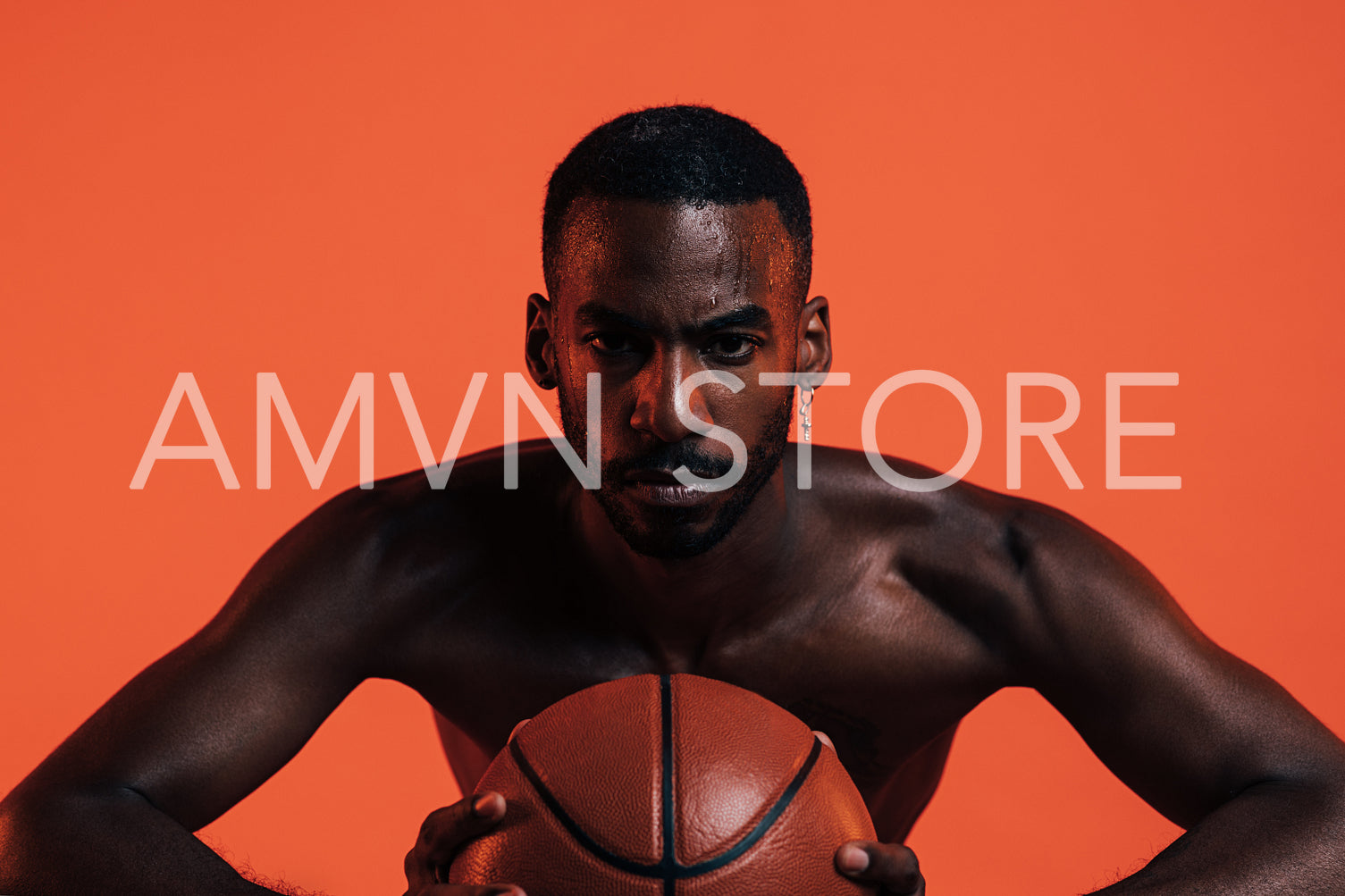 Bare-chested sportsman clutching basket ball in front of him while standing in a studio