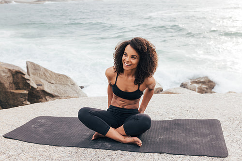 Smiling woman sitting on a mat looking away. Muscular female in fitness wear taking a break by ocean.