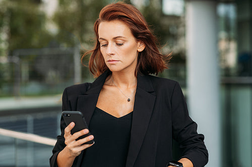 Stylish businesswoman with ginger hair wearing black formal clothes looking at mobile phone