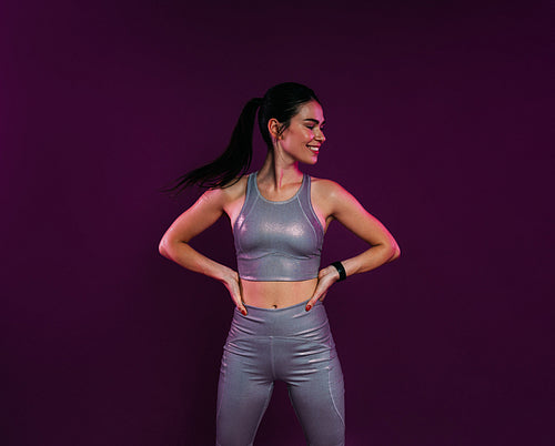 Young woman in silver fitness attire with hands on hips looking away at magenta background