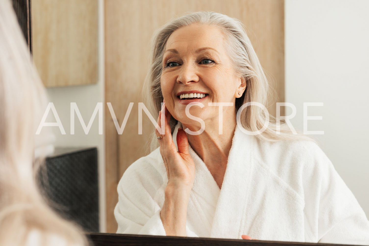 Happy aged female touching her face in front of a mirror. Woman in bathrobe admires her reflection.