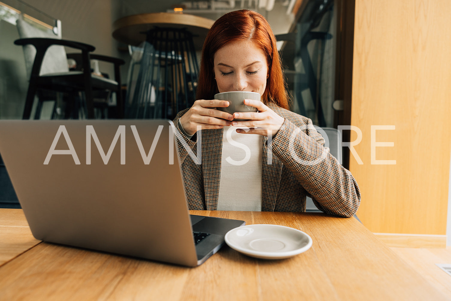 Smiling woman with ginger hair enjoying her morning coffee. Businesswoman holding a cup with closed eyes while sitting at a table.