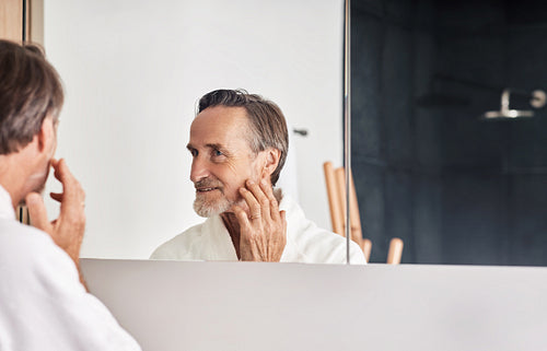 Smiling senior man looking at his reflection in the mirror and touching face. Mature male in a bathroom in the morning.