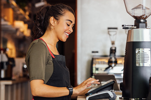 Side view of smiling waitress in apron typing digital cash terminal in coffee shop