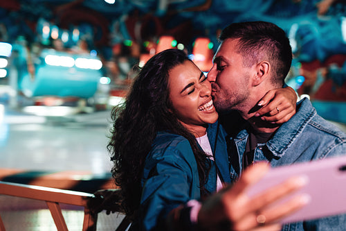 Boyfriend kissing his girlfriend while she is making selfie. Young couple during a summer festival.