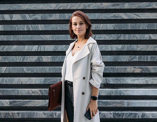 Stylish middle-aged female entrepreneur holding leather folder standing outdoors