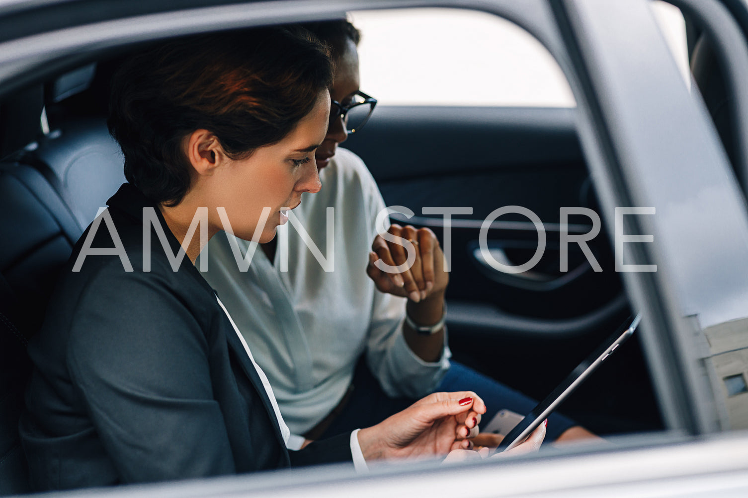 Two female colleagues looking at digital tablet and talking while sitting of a backseat of a taxi	