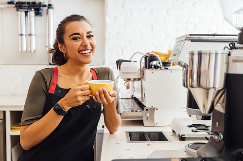 Happy female barista standing at coffee machine and holding a mug. Female cafe worker taking a break.