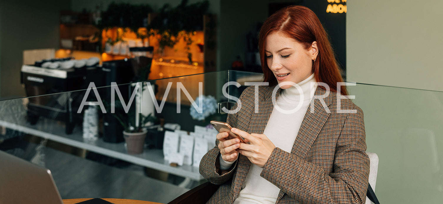 Redhead woman in formal wear sitting in a cafe holding a mobile phone and smiling