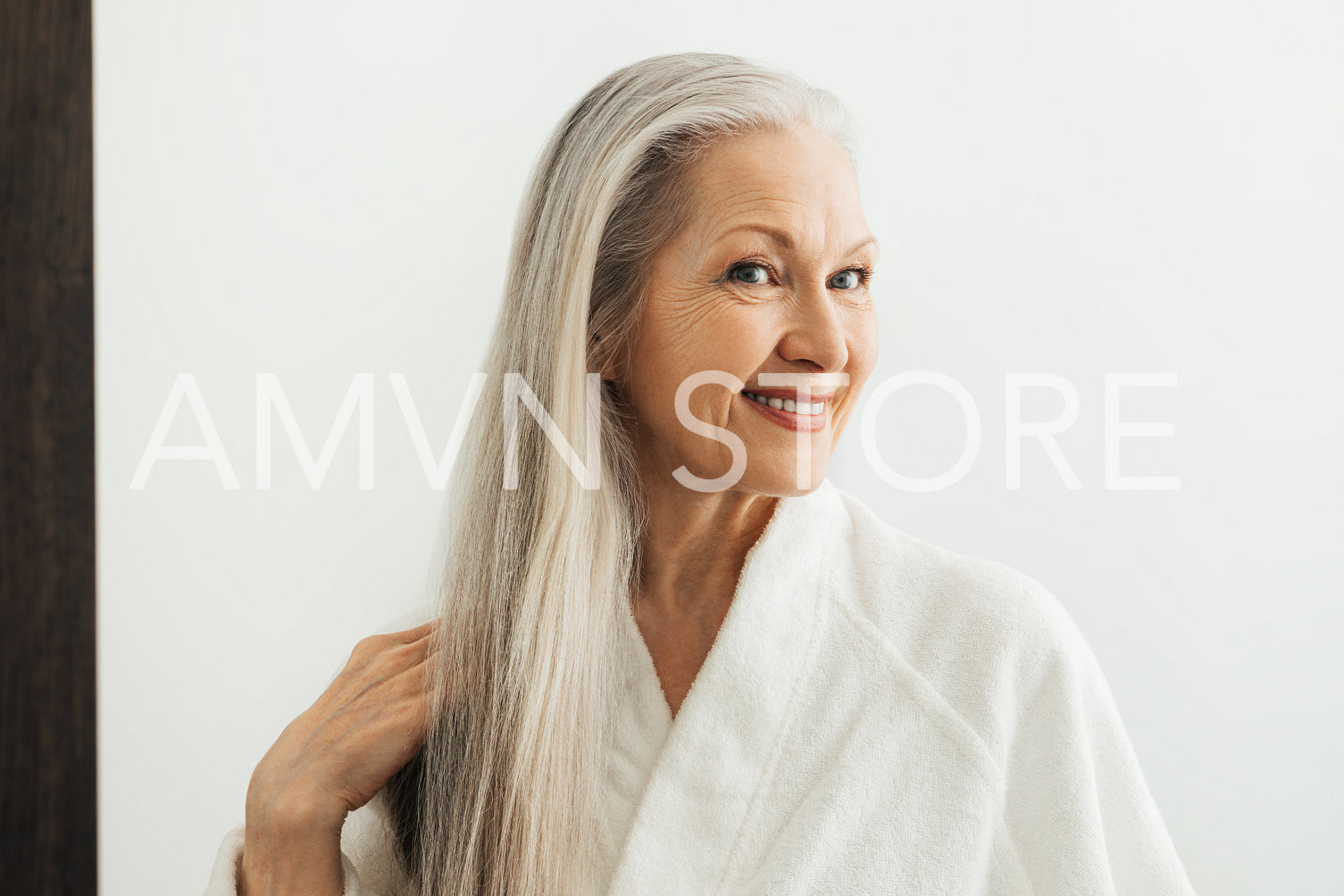 Senior female with grey hair in a bathrobe looking at a mirror in a bathroom. Woman adjusting her grey hair at morning.