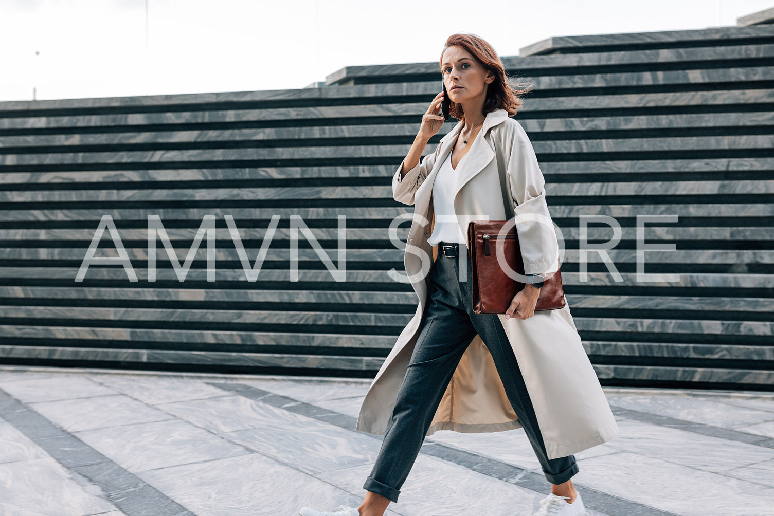 Stylish woman in a coat walking outdoors talking on a mobile phone. Businesswoman with ginger hair and a leather folder walking on the street.