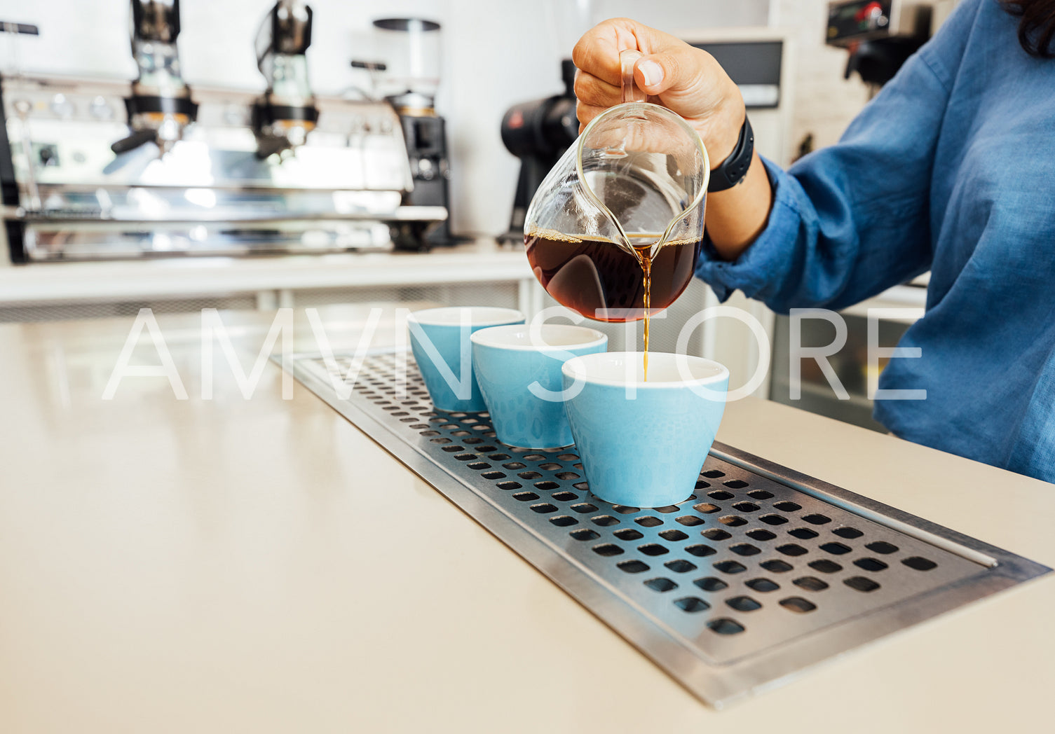 Hand of a female barista holding a glass teapot and pouring coffee into a small blue cup	