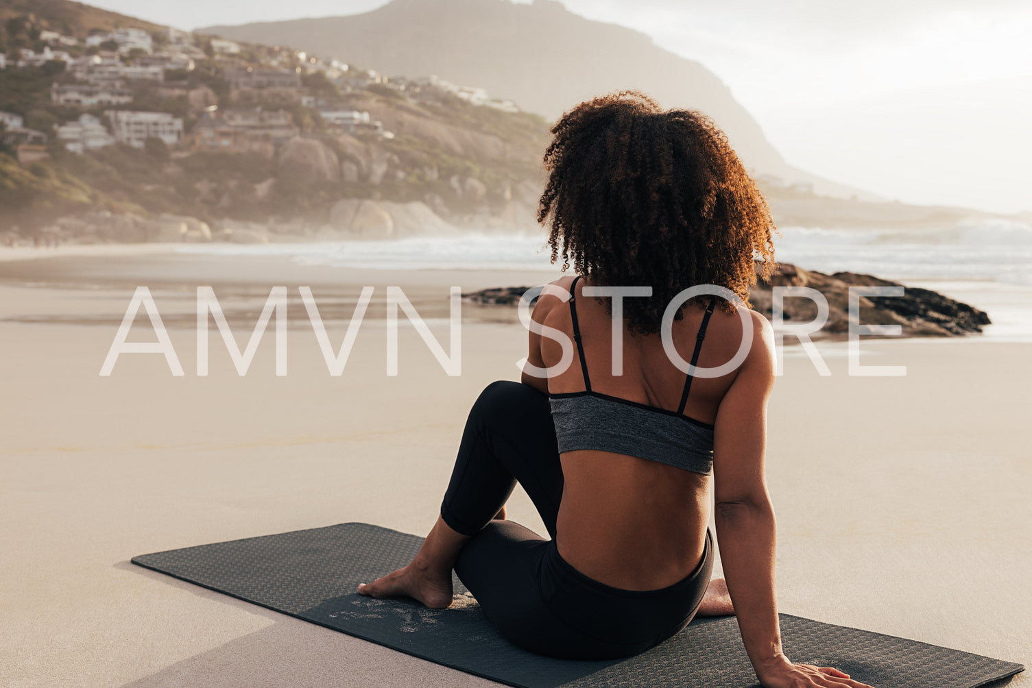 Back view of a young female practicing yoga at sunset. Woman in fitness wear sitting on a mat looking at hills on a shore.