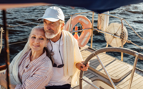 Mature couple sitting on yacht and wrapped in blanket. Elderly people enjoying a vacation on sailboat.