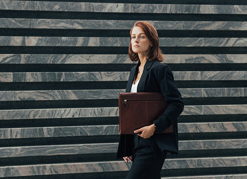 Confident middle-aged female in formal black clothes walks outdoors with a leather folder. Female with ginger hair is going to a corporate meeting.