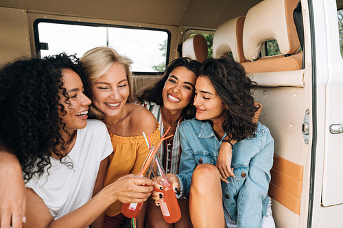 Two smiling women toasting with bottles in van. Close friends enjoying summer road trip.