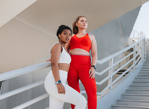 Two plus size females in sportswear standing on a staircase. Young women relaxing after exercises outdoors.