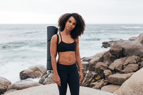 Portrait of a beautiful fit woman with yoga mat looking at camera while standing outdoors by coast