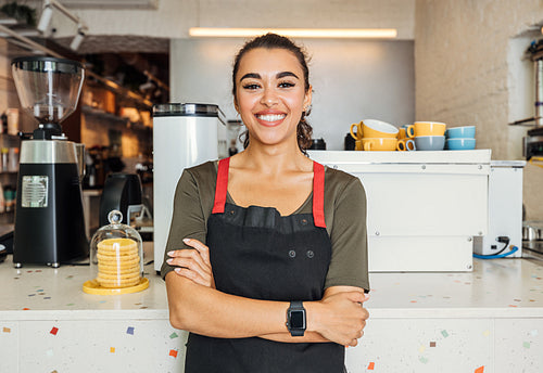 Female barista standing arms crossed in a cafe. Smiling waitress looking at camera while standing in front of a counter.